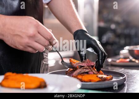 Nahaufnahme eines unerkennbaren Küchenchefs in Handschuhen, der Fleischsteak auf dem Teller mit Gemüse in der kommerziellen Küche zugibt Stockfoto