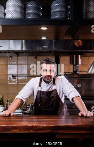 Porträt des selbstbewussten reifen bärtigen Koch des Restaurants mit Lederschürze lehnte sich auf Holztisch Stockfoto