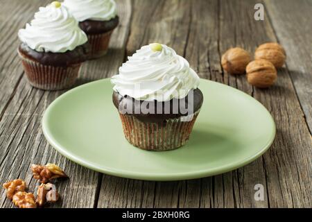 Schokoladenkuchen mit Schlagsahne. Vor dem Teller liegen Walnusskerne, zwei Cupcakes und Walnussschalen in verschwommenem Hintergrund. Stockfoto