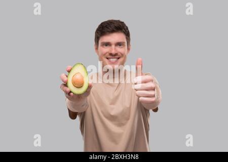 Mann zeigt Avocado und Daumen nach oben Nahaufnahme isoliert. Avocado in zwei Hälften geschnitten Stockfoto