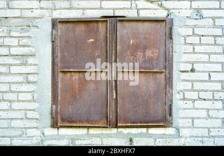 Fensteröffnung zum Entladen von Waren im Laden, geschlossen durch rostige Eisenläden an der weißen Ziegelwand. Teilweise mit Eisenläden verschlammte Stockfoto