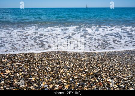 Schäumende Welle am ruhigen Strand. Ufer mit Kieselsteinen. Blauer Himmel Seeseite mit Segelboot am Horizont. Stockfoto