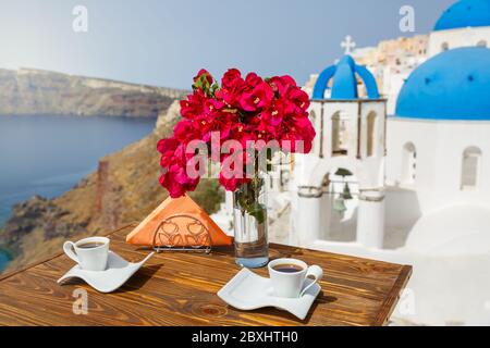 Zwei Tassen Kaffee und ein Zweig blühender Bougainvillea vor der Kulisse der Architektur der Insel Santorini Stockfoto