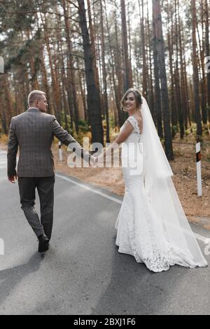 Lächelnde Braut und Bräutigam gehen auf der Straße im Wald von hinten fotografiert. Porträt von Braut und Bräutigam Stockfoto