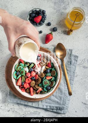 Trendige Speisen - Pfannkuchen Müsli. Milch auf Haufen von mehrfarbigen Mini-Cerealien Pfannkuchen in Schüssel gießen. Winzige Cerealien-Pfannkuchen mit Beeren in Bastelplatte auf grauem Zement Hintergrund. Vertikal. Draufsicht, flaches Lay Stockfoto