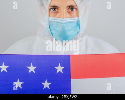 Porträt einer kaukasischen Ärztin mit psa-Anzug und Maske im Krankenhaus. Hält die Flagge der Vereinigten Staaten. Frau vor dem Coronavirus geschützt. Stockfoto