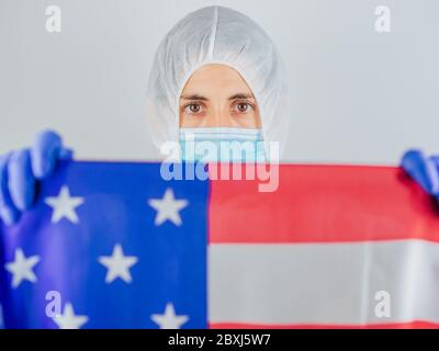 Porträt einer kaukasischen Ärztin mit psa-Anzug und Maske im Krankenhaus. Hält die Flagge der Vereinigten Staaten. Frau vor dem Coronavirus geschützt. Stockfoto