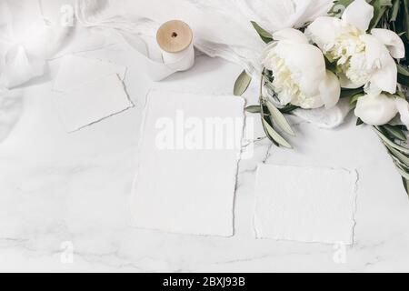 Hochzeitspapier, Stillleben Komposition. Grußkarten Mockup Szene. Bouquet von weißen Pfingstrosen Blumen, Olivenzweige, Kuh Petersilie auf Marmor, Seide Stockfoto