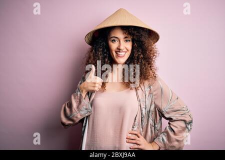 Junge schöne Frau mit lockigen Haaren und Piercing trägt traditionelle asiatische konische Hut tun glücklich Daumen hoch Geste mit der Hand. Ausdruck wird genehmigt Stockfoto