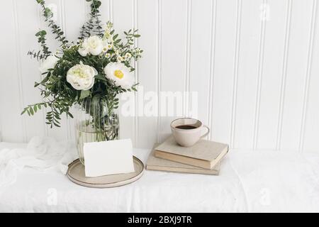 Feminine Hochzeit oder Geburtstagstisch Mockup Szene mit Blumenstrauß. Weiße Pfingstrosen, tanacetum-Blumen, Eukalyptus, Grußkarte, Tasse Kaffee und Buh Stockfoto