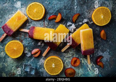Hausgemachte Orangen- und Pflaumenpopsicles auf grauem Hintergrund, Draufsicht mit Pflaumen- und Orangenstücken. Stockfoto