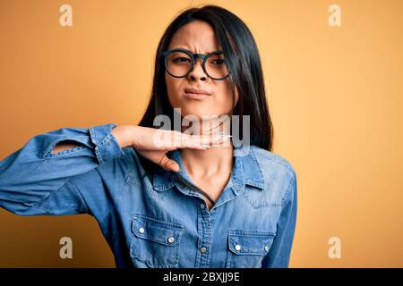 Junge schöne chinesische Frau mit lässigen Denim-Shirt über isolierten gelben Hintergrund Schneiden Kehle mit der Hand als Messer, bedrohen Aggression mit f Stockfoto