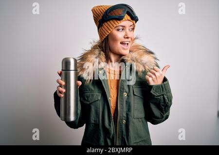 Junge schöne Skifahrer Frau mit blauen Augen tragen Skibrille trinken Thermo mit Kaffee zeigen und mit dem Daumen bis zur Seite mit glücklichen f zeigen Stockfoto