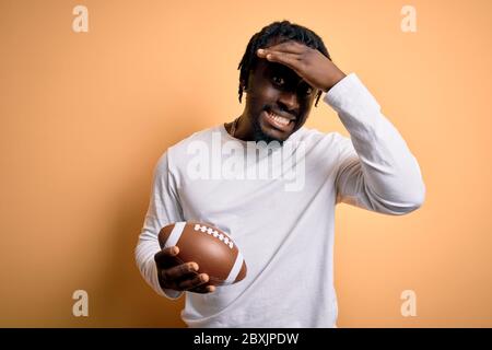 Junger afrikanischer Spieler Mann, der Rugby spielt, der American Football Ball über gelbem Hintergrund hält, mit Hand auf Kopf gestresst, schockiert von Scham und Surpri Stockfoto