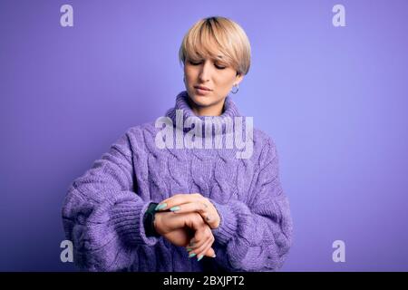 Junge blonde Frau mit kurzen Haaren trägt Winter Rollkragen Pullover über lila Hintergrund Überprüfung der Zeit auf Armbanduhr, entspannt und zuversichtlich Stockfoto