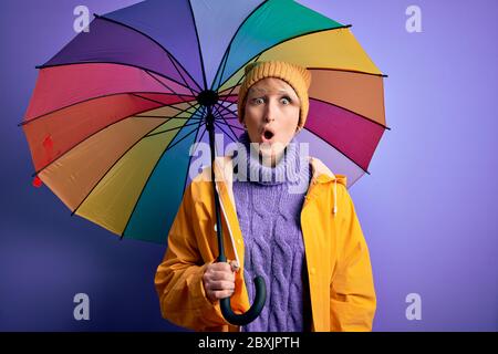 Junge blonde Frau mit kurzen Haaren in wasserprof Regenmantel und bunten Regenschirm erschreckt im Schock mit einem Überraschungsgesicht, Angst und aufgeregt mit Fea Stockfoto