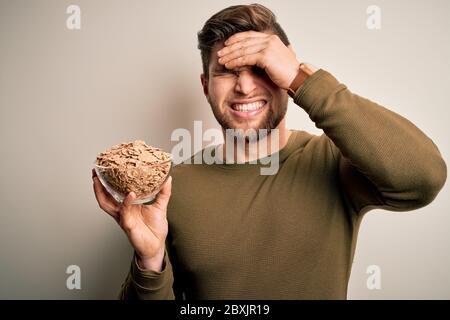 Junge blonde Mann mit Bart und blauen Augen hält Schüssel mit Cornflakes Getreide gestresst mit Hand auf den Kopf, schockiert mit Scham und Überraschung Gesicht, wütend Stockfoto