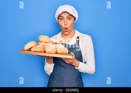 Junge schöne blonde Bäckerin Frau mit blauen Augen hält Tablett mit hausgemachten frischen Brot erschrocken im Schock mit einer Überraschung Gesicht, Angst und aufgeregt mit Stockfoto
