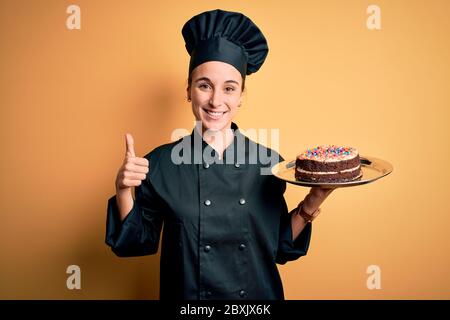 Junge schöne Bäckerin Frau trägt Herd Uniform und Hut halten Tablett mit Kuchen glücklich mit großem Lächeln tun ok Zeichen, Daumen hoch mit den Fingern, ausgezeichnet Stockfoto