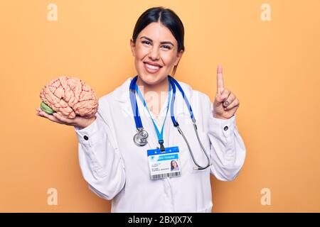Junge schöne Psychiaterin Frau trägt Stethoskop halten Gehirn über gelben Hintergrund lächelnd mit einer Idee oder Frage Zeigefinger mit Happ Stockfoto