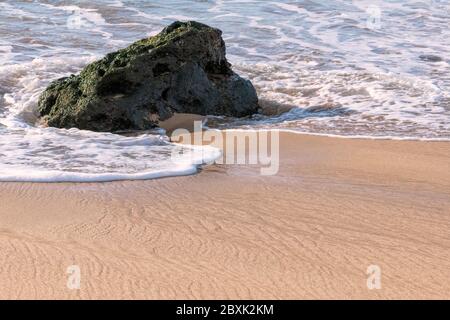 Eine Welle umhüllt ein Stück Lavagestein an einem Sandstrand. Aufnahme am Papohaku Beach, Molokai, Hawaii. Stockfoto