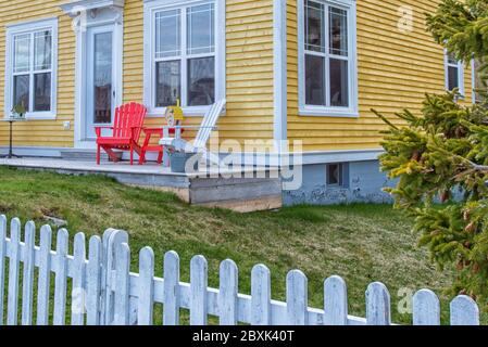 Eckansicht eines Vintage gelben Holzklatschtafelhauses mit roten und weißen Adirondack-Stühlen auf der Terrasse. Das Gebäude hat mehrere Fenster. Stockfoto