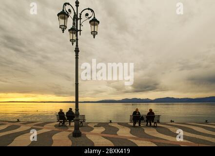 Landschaftlich schöner Blick auf das Ufer des Gardasees mit Menschen sitzen auf Bänken auf der Promenade bei Sonnenuntergang, Lazise, Verona, Venetien, Italien Stockfoto