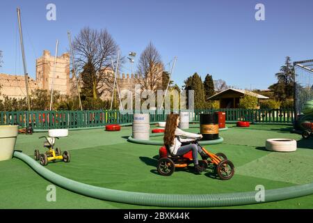 Langhaarige kleine Mädchen (9 Jahre), die auf einem Tretauto auf einem Kinderspielplatz vor der Scaliger Burg, Lazise, Verona, Venetien, Italien, reitet Stockfoto