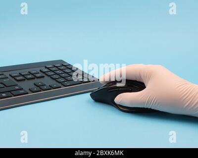 Hand in Gummihandschuh hält eine Maus in der Nähe der Tastatur auf einem blauen Hintergrund. Stockfoto