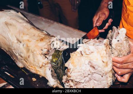 Das gebratene Schwein auf dem Tisch, das als "Lechon" bekannt ist, mit einem Metzgermesser mit der aromatischen Zitronengras-Füllung von Hand schneiden. Selektiver Fokus. Stockfoto