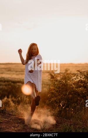 Junge schöne Frau, die mit dem Fuß Staub macht und an einem sonnigen Sommertag im Weizenorangenfeld schreit. Verrückt. Sich frei und glücklich fühlen. Mi Stockfoto