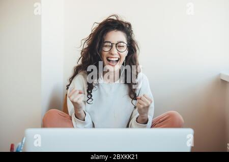 Schöne junge Frau mit Brille und lockiges Haar lächelnd und gestikulierenden die gewinnen Emotionen vor dem Laptop zu Hause Stockfoto