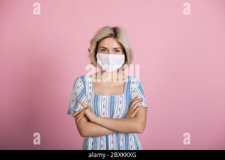 Blonde kaukasische Frau, die eine medizinische Maske und ein Kleid trägt, schaut auf die Kamera auf einer rosa Studiowand Stockfoto
