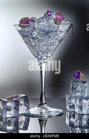 Weißgold Schmuck mit Amethysten und blauem Topas in einem martini-Glas mit Eis und Eiswürfeln auf grauem Hintergrund mit Spiegelung. Schmuck Kunst und produ Stockfoto