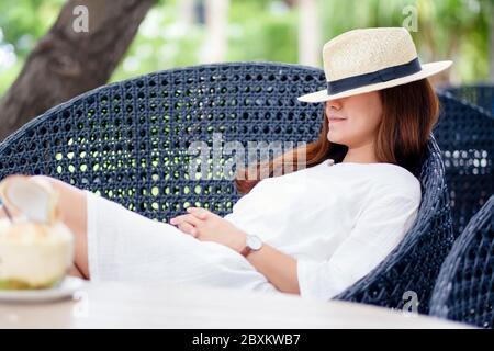 Porträtbild einer schönen asiatischen Frau mit Hut liegend und schlafen auf einer Bank im Garten mit Kokosnuss auf dem Tisch Stockfoto