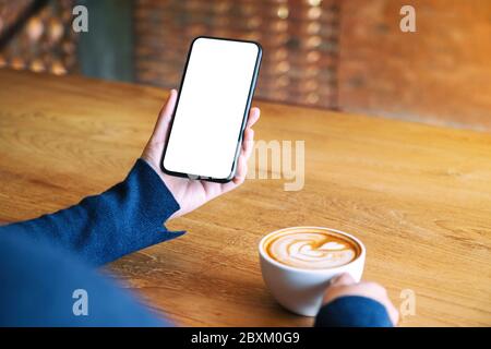 Mockup-Bild einer Frau, die weißes Mobiltelefon mit leerem Bildschirm hält, während sie Kaffee auf einem Holztisch im Café trinkt Stockfoto