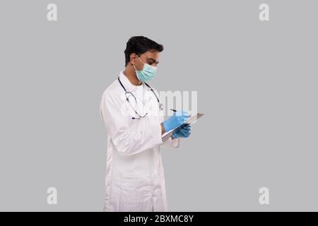 Doktor, der in der Zwischenablage schreibt, trägt medizinische Maske und Handschuhe. Indischer Mann Doktor Zwischenablage Isoliert Stockfoto