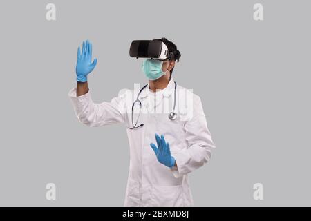 Arzt trägt VR-Brille, medizinische Maske und Handschuhe Hände an den Seiten isoliert. Indian man Doctor zeigt in der virtuellen Realität Stockfoto