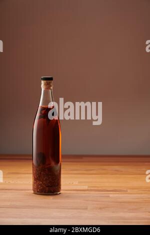 Transparente Flasche hausgemachtes Chilliöl mit Stücken von trockenem Chili, auf einem Holztisch Stockfoto