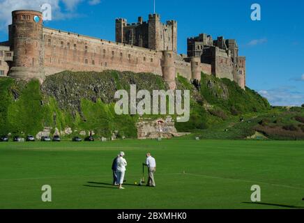 Drei Senioren spielen Krocket auf dem Rasen vor der imposanten Kulisse von Bamburgh Castle, Northumberland, Großbritannien Stockfoto