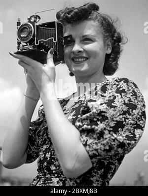 Amateurfotograf in den 1940er Jahren. Eine junge Frau fotografiert an einem Sommertag. Das Kameramodell ist praktisch. Wenn Sie es nicht verwenden, werden die Linse und der Balg in das Kameragehäuse gefaltet. Um Bilder aufzunehmen, haben Sie es geöffnet und waren bereit zu gehen. Die Kamera produzierte analogen Film. Stockfoto