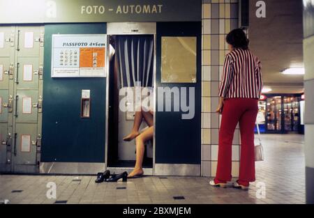 In den 1970er Jahren. Ein Fotostand an einer U-Bahn-Station, die durch das Aussehen von es für andere Fotografie dann die üblichen Passfotos verwendet wird. Eine Frau steht und schaut auf die Person, die ihre Schuhe entfernt hat und sitzt beiläufig drinnen, um ihr Foto aufzunehmen. Schweden 1971. ref CV48-5 Stockfoto