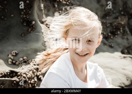 Ein junges Mädchen sitzt an einer Strandklippe mit Seepocken im Hintergrund, ihre blonde blonde Haare ist windgeblasen und sie schaut auf die Kamera, ein genommen