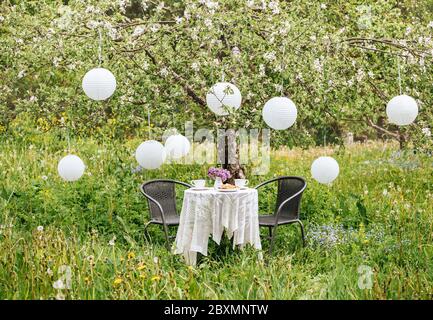 Niedliches Arrangement mit Gartenmöbeln, zwei Teetassen und Kuchen auf dem Tisch. Viele chinesische Papierlaternen hängen vom blühenden Apfelbaum zur Dekoration. Stockfoto