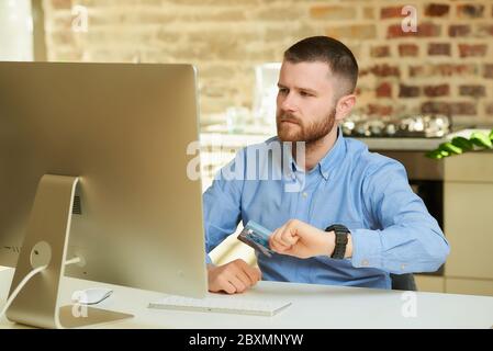 Ein Mann mit Bart sieht sich das Display des Desktop-Computers an und hält eine Kreditkarte zu Hause. Ein Kerl wartet ungeduldig, um eine Online pa tun Stockfoto