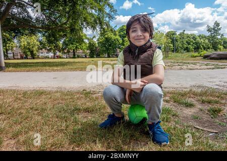 Ein kleiner Junge sitzt auf einem Fußball in der Par, posiert und schaut in die Kamera und lächelt. Es ist windig und seine Haare flattern in der Brise. Stockfoto