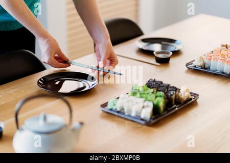 Frauenhände arrangieren den Tisch mit verschiedenen Sushi-Sets auf einem Holztisch in einem sonnigen Zuhause Stockfoto