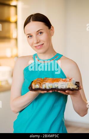 Porträt charismatische Frau präsentiert einen Teller mit Sushi-Rollen in den Händen mit moderner Küche im Hintergrund Stockfoto