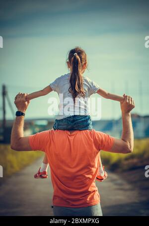 Papa tanzt auf seinen Schultern mit seiner Tochter in der Sonne. Vater reist mit Baby auf seinen Schultern in Strahlen des Sonnenuntergangs. Kind mit Eltern spaziert bei Sonnenuntergang Stockfoto