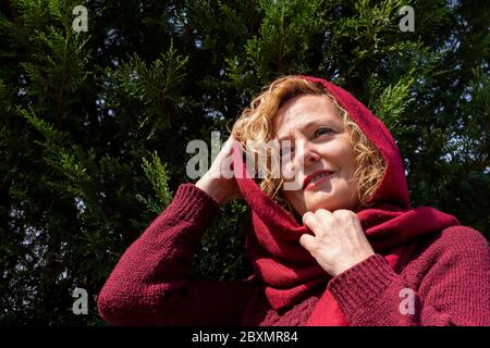 Eine blonde Frau mittleren Alters spielt die Rolle der kleinen roten Reithaube wie im berühmten Märchen, gegen Thuja-Bäume Stockfoto